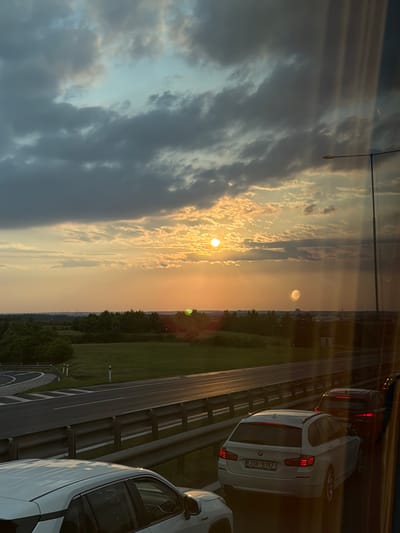 The image depicts a sunset scene on a highway, likely in or near Kuří, Czechia. The sky is filled with dramatic, dark clouds interspersed with patches of bright orange and yellow light from the setting sun. The sun itself is visible as a bright orb near the center of the horizon.
In the foreground, several cars are visible on the highway. A white BMW station wagon with the license plate "2SN 5157" is in the right lane, and a red SUV is behind it. To the left, the roof of a white vehicle is partially visible. The highway itself has multiple lanes separated by metal guardrails. The road surface is wet and reflects the sunset light.
Beyond the highway, there is a stretch of green fields and trees, with a winding road and some white posts visible on the left side of the frame. In the distance, a line of trees forms the horizon, and a few faint structures can be seen. A tall lamppost stands on the right side of the highway, partially obscuring the sky. Vertical streaks of light, possibly lens flares or reflections, are visible on the right side of the image. The overall atmosphere is calm and somewhat moody due to the heavy clouds and sunset lighting.