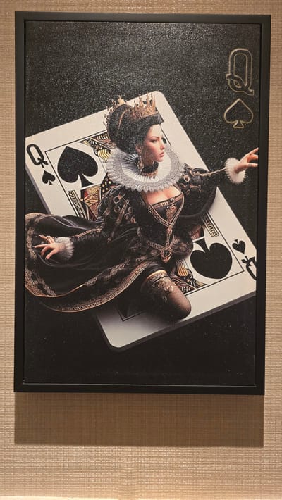 The image depicts a framed artwork featuring a stylized depiction of the Queen of Spades. The queen is shown in a regal, ornate costume with a crown, ruff collar, and detailed fabric. She is positioned on top of a large playing card, the Queen of Spades, which is itself set against a dark, textured background with glitter or sparkle accents. The artwork is hanging on a textured wall, possibly wallpapered, in a neutral beige or light brown color. The lighting suggests an indoor setting, with a shadow cast on the wall below the frame. The specific location in Agios Epiktitos, Northern Cyprus / Cyprus is not discernible from the visual information provided in the image.