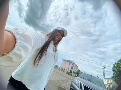 A low-angle, wide-angle selfie taken outdoors on an overcast day shows a young woman from the chest up, facing the camera with a slight smile. She is wearing a white baseball cap and a crinkled white shirt with elastic at the cuffs. Her long, dark hair is visible. In the background, a gray SUV is parked. Beyond the vehicle, a pink and green building with several windows is visible, along with utility poles and wires. The ground appears to be a mixture of asphalt and gravel. The scene is consistent with an outdoor urban environment in Akyar, Russia.