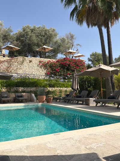 This image depicts a tranquil outdoor swimming pool area, likely a resort or private villa, bathed in bright sunlight. The pool itself is a vibrant turquoise, with visible "200" marking on its edge. The surrounding patio is paved with light-colored stone tiles.

Several lounge chairs, upholstered in dark fabric with striped cushions, are arranged on the patio. Some chairs have small wooden tables between them. Two large potted plants, one terracotta and one a darker shade, are placed near the chairs. In the background, a tiered stone wall supports lush greenery, including vibrant pink flowering bushes and tall palm trees. Several light-colored umbrellas are positioned around the area, offering shade. A glimpse of two people can be seen in the far left background, near some of the umbrellas.

The overall atmosphere is one of relaxation and luxurious leisure, set against a backdrop of natural beauty and manicured landscaping. The location is identified as the city of Montuïri, Spain, suggesting a Mediterranean setting.