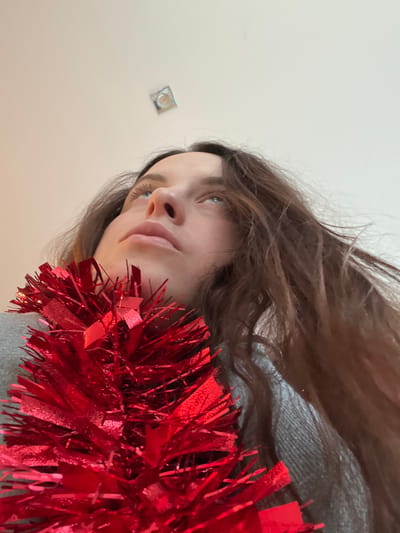 A low-angle, upward-looking photograph depicts an adult female subject indoors. The subject has long, dark brown hair and light-colored eyes, gazing directly upwards towards a plain white ceiling. A dense, shimmering red tinsel garland is draped across the subject's upper chest and neck, partially obscuring the lower face. The subject wears a grey garment, visible beneath the tinsel. In the upper-center of the white ceiling, a single, recessed light fixture, appearing square or circular, is visible. The specific location in the city of Callbach, Germany, or any other geographic detail beyond being indoors, cannot be determined from the visual information provided.