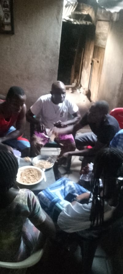 A group of people sit around a small table indoors, sharing food from two dishes. The setting appears to be a modest home with simple furnishings and walls. The participants are dressed casually. Lighting is dim. The subjects of the photo seem to be involved in a social gathering, possibly sharing a meal or having a conversation.