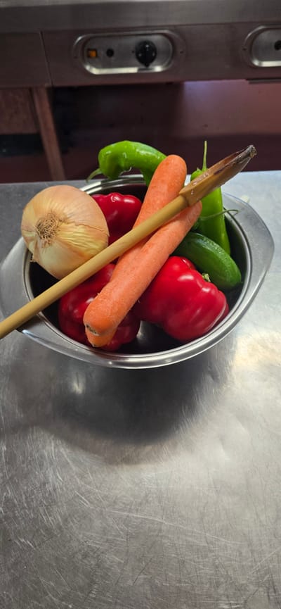 A close-up, horizontal, eye-level shot showcases a stainless steel bowl filled with fresh vegetables. The bowl is positioned on a scratched stainless steel surface, possibly a countertop or table. Inside the bowl, a mix of red bell peppers, green chili peppers, and carrots are visible. A prominent wooden utensil, possibly a spoon or spatula, lies diagonally across the vegetables. The wooden utensil appears worn, with some charring at the top. In the foreground, a round, light-brown onion rests at the bottom of the bowl. The overall lighting is bright, creating reflections on the metallic surfaces and highlighting the vibrant colors of the produce. The background is slightly out of focus, revealing a hint of a dark, reddish-brown surface and a stainless steel appliance with control knobs. The scene suggests food preparation or ingredients ready for cooking. There is no visible text.