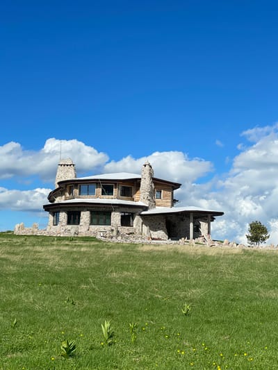 The image shows a distinctive, multi-story stone and wood house situated on a grassy hilltop under a bright blue sky with scattered white clouds. The house has a circular or polygonal shape, with multiple windows and two prominent stone chimneys. The lower level appears to be constructed primarily of rough-hewn stones, while the upper level incorporates wooden elements and more finished windows. A low stone wall surrounds the immediate area of the house, and a single tree stands to the right. The foreground is a field of green grass, with some smaller yellow wildflowers visible. The scene depicts a rural or semi-rural setting with a unique architectural style. The provided information about the city of Gela, Bulgaria is not verifiable from the image itself.