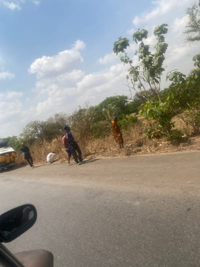 The scene is located in the city of Ninzo West, Nigeria. The image displays a section of a grey asphalt road under a light blue sky with scattered white cumulus clouds. On the far left, a white vehicle is partially visible, equipped with multiple yellow jerrycans secured to its roof rack. Five individuals are present along the right side of the road and its adjacent dry, dirt shoulder. From left to right: one individual in dark clothing stands near the white vehicle; a second individual in light-colored clothing crouches on the roadside; a third individual in a blue striped shirt stands upright on the road; a fourth individual, partially obscured behind the third, wears a red garment; and a fifth individual, dressed in an orange-brown full-body garment, stands on the dirt shoulder, facing away from the camera. The environment consists of dry, light brown grasses and scattered green-leafed trees and bushes lining the roadside. The lower-left corner captures a partial view of a vehicle's side mirror and an occupant's arm.
