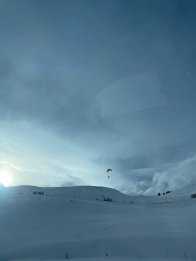 This image depicts a snow-covered mountainous landscape in the vicinity of Le Monêtier-les-Bains, France, under a cloudy sky. A single individual is engaged in paragliding, visible in the mid-right portion of the frame. The paraglider features a bright yellowish-green canopy, with the pilot suspended beneath it.
The foreground and midground consist of extensive, undulating terrain completely covered in white snow, indicating a winter environment. Patches of dark, bare vegetation or small trees are discernible on the snow-covered slopes in the midground, particularly on the left side, and some evergreen trees are visible on a distant slope to the right.
The sky occupies approximately the upper two-thirds of the image, characterized by a gradient of light blue and grey clouds. A bright, localized glare from the sun is present on the far left horizon, partially obscured by clouds. Subtle, irregular reflections are visible across the upper portion of the image, indicative of the photograph being taken through a glass surface. The overall lighting is soft and diffuse, consistent with an overcast day.