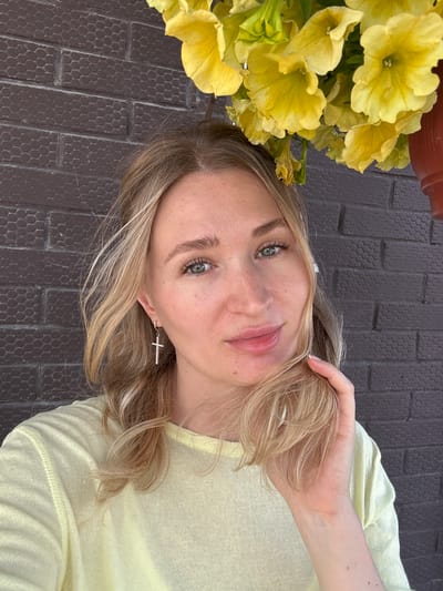 A blonde woman with blue eyes is pictured in a close-up shot against a dark brick wall. She is wearing a light yellow long-sleeved shirt and a silver cross earring. Her right hand is holding a section of her hair near her face, and she is looking directly at the camera. Above her head, a cluster of bright yellow flowers is visible, slightly out of focus. The overall lighting suggests an outdoor, daytime setting.