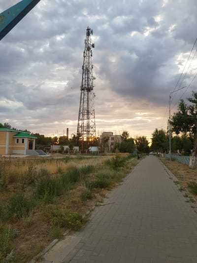 This media file depicts an outdoor scene, likely in Kurchatov, Kazakhstan, during the late afternoon or early evening. The sky is dominated by a dramatic display of clouds, with patches of sunlight breaking through, suggesting a partly cloudy or clearing weather condition. The sun is low on the horizon, casting a warm, golden light across the scene.
The central object is a tall, lattice telecommunications tower, adorned with antennas and dishes. To the right of the tower, a paved walkway stretches into the distance, bordered by dry, grassy vegetation. In the background, several buildings are visible, including a brick structure with a green roof and what appears to be industrial elements like a smokestack. Trees line the path and the buildings, their leaves catching the warm light. A metal fence with a blue railing can be seen along the edge of the walkway.
There are no people visible in the image, and the scene conveys a sense of quietude and natural beauty, enhanced by the atmospheric lighting. The overall impression is one of a peaceful, somewhat utilitarian, urban or semi-urban environment at dusk. No visible text is present.