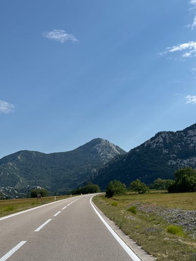 The image captures a scenic mountain road on a clear, sunny day in Dragalj, Montenegro. A paved asphalt road with white lane markings curves through a grassy field and leads towards a backdrop of lush green mountains. The mountains are densely covered in trees and feature rocky outcrops, with one prominent peak in the center. Scattered clouds drift across the bright blue sky. The roadside is lined with grass, small wildflowers, and a rocky embankment. There are no people or vehicles visible in the image, suggesting a tranquil and unpopulated setting. A small signpost is visible on the left side of the road, near the edge of the field.