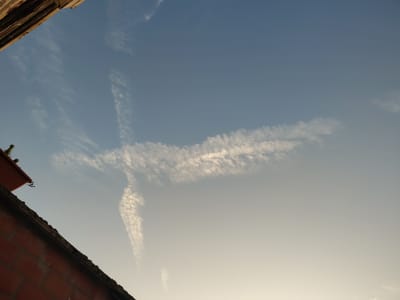 This media file shows a daytime view of a sky with wispy clouds. The clouds are arranged in a way that resembles a cross shape. The sky is a clear blue, gradually fading to a lighter hue towards the horizon.
In the foreground, the top of a brick wall and a section of a roof are visible, suggesting an outdoor setting with buildings. The texture of the bricks is detailed, and the roof appears to be made of orange tiles. A hint of greenery, possibly a plant or tree, can be seen near the top left corner, peeking over the roofline.
The lighting indicates it is either early morning or late afternoon, given the soft gradient of the sky and the shadows that might be cast but are not prominently featured. The weather appears to be fair, with the presence of light, scattered clouds. The location is identified as Nederbrakel, Belgium, implying a European setting. There are no visible people or any text in the image. The overall impression is serene, with the cloud formation being the most striking element.