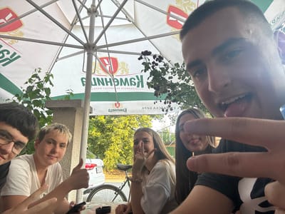 The media file is a low-angle, wide shot taken outdoors on a sunny day. The scene features a group of five young people, likely friends, gathered around a table under a large white umbrella with red and green branding. The umbrella has the text "KOWENNG" visible multiple times.
The people are positioned around the umbrella, with some in the foreground and others in the background. One person in the foreground is sticking out their tongue and making a peace sign with their fingers, suggesting a lighthearted and playful atmosphere. Another person is making a "rock on" hand gesture. They appear to be taking a selfie or having their photo taken in a casual setting.
In the background, there's a white car and a bicycle, with lush green trees and foliage. The architecture suggests a building or outdoor establishment. The bright sunlight and green foliage indicate it's likely daytime during a warm season. The overall mood seems cheerful and relaxed.
The text "KOWENNG" is prominently displayed on the umbrella. There is also partially visible text on banners in the background, but it is not clearly legible. The location context of Novi Chiflik, Bulgaria, suggests they might be at an outdoor cafe, restaurant, or recreational area.