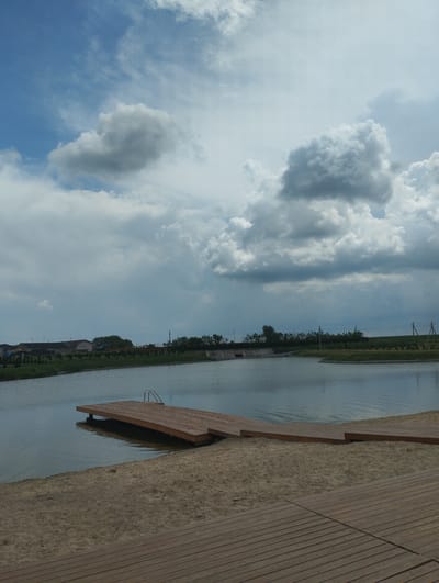 This media file depicts an outdoor scene with a lake, a wooden pier, and a sandy beach. In the background, a line of trees and buildings can be seen along the opposite shore. The sky is filled with dramatic, dark clouds, suggesting impending or recent rain. The water in the lake is calm and reflects the sky. A wooden pier extends from the sandy beach into the lake, with metal steps leading down into the water. The overall mood is serene and somewhat moody due to the cloudy sky. The time of day appears to be late afternoon or early evening. There is no visible text in the media file. The location is indicated as Romanivka, Ukraine.