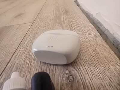 A white wireless earbud charging case with the JBL logo is centered on a light brown wooden floor with a visible grain pattern. Three small LED lights are illuminated on the front of the case. To the left of the case, a white bottle cap is visible. To the right and slightly below the case, a black object is partially visible. The floor meets a rough, gray wall with some damage along the baseboard. The scene appears to be indoors, likely in a residential setting. There is no indication of the location being in Avtury, Russia.