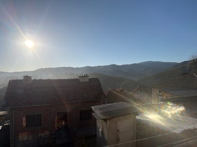 The image depicts a view in the village of Grashevo, Bulgaria. It's a sunny day, and the scene includes houses with red-tiled roofs and a small concrete outbuilding with a door. In the background, there are hills or mountains partially obscured by a light haze. A chimney is visible on the nearest house. The strong sunlight causes lens flare in the lower right portion of the image.