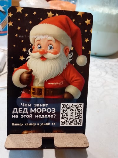 This is a close-up, eye-level shot of a wooden phone stand with a printed insert featuring a cartoon depiction of Ded Moroz (Grandfather Frost), a character similar to Santa Claus. Ded Moroz is shown from the chest up, wearing his iconic red hat with white trim, a red coat, and a large, golden belt buckle. He has a white beard and mustache, blue eyes, and rosy cheeks, and is giving a thumbs-up. The background behind Ded Moroz is dark blue with gold stars.

Below the illustration of Ded Moroz, there is text in Russian. The first line reads "Чем занят" (What is occupied with). The second line reads "ДЕД МОРОЗ" (GRANDFATHER FROST). The third line asks "на этой неделе?" (this week?). Below this, there is an instruction: "Наведи камеру и узнай! >>" (Point your camera and find out! >>).

To the right of the text is a QR code. Below the QR code, there is a small number "1124744". In the lower part of the image, the wooden phone stand itself is visible, designed to hold a phone upright. The scene appears to be indoors, possibly on a table covered with a light-colored tablecloth. The overall mood is festive and playful, related to the winter holiday season. The location context is Докшыцкі сельскі Савет, Belarus.