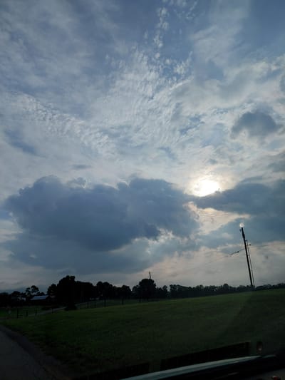 The image depicts a cloudy sky over a rural landscape in Stamping Ground, United States. The sun is partially visible through a gap in the dark, heavy clouds, casting a bright glow. The foreground features a lush green field, with trees and a few buildings forming a dark silhouette against the horizon. A utility pole with wires is visible on the right side of the image. The overall scene suggests an overcast day, possibly before or after a storm.