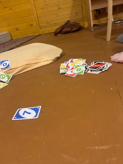 The image shows a floor with several UNO cards scattered on it. There is a beige blanket or cushion on the left side of the image, and a pile of UNO cards in the center. A few more UNO cards are visible in the foreground and on the right side. In the background, there are wooden walls and part of a wooden table. A person's foot is visible on the far right of the image. The overall setting appears to be an indoor space, possibly a room in a house. The city of Барское-Городище, Russia, is not visible or indicated in the image.