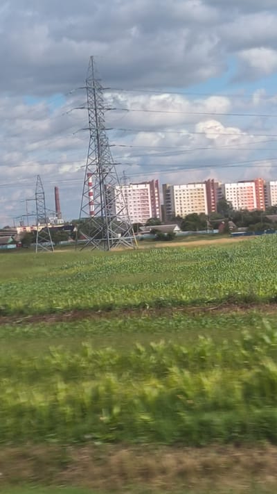 The image depicts a rural landscape with industrial elements and residential buildings in the distance. A large, lattice-style electricity pylon dominates the left side of the frame, with power lines extending horizontally across the sky. To the left of the pylon, a red smokestack from an industrial facility is visible, along with other structures of similar construction.
In the mid-ground, a field of green crops, possibly corn, stretches across the scene. Behind the field, a row of multi-story apartment buildings with red and white facades are situated. These buildings appear to be of a Soviet-era architectural style. Some smaller, single-story houses are scattered between the industrial area and the apartment buildings.
The sky is filled with dramatic, heavy clouds, suggesting a partly cloudy or overcast day. The foreground of the image shows a blurred effect, indicating movement, possibly captured from a vehicle. The overall scene suggests a location where agricultural land meets industrial development and residential areas, characteristic of peri-urban or rural settings near industrial centers. The location is identified as Krasnenski sielski Saviet, Belarus.