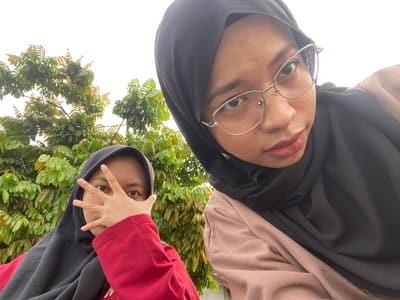 Two young women, likely teenagers, are captured in a selfie outdoors, with lush green foliage and a white sky forming the background. The woman on the right, closer to the camera, wears a black hijab and glasses with thin silver frames. Her face, turned slightly towards the viewer, displays a neutral expression. She is also wearing a light brown jacket over a dark garment. The woman on the left, partially obscured by her hand, also wears a black hijab and a red top. Her left hand, with fingers spread, is held up in front of her face, obscuring her eyes. She is positioned behind and to the left of the other woman. The overall setting suggests a casual outdoor gathering, possibly in a park or residential area, consistent with locations in Air Molek, Indonesia.