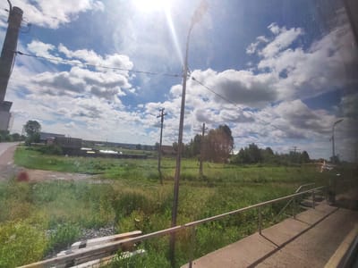 This image, taken on a bright, sunny day, captures a rural landscape in Malaya Bod'ya, Russia. The sky is a vibrant blue, filled with fluffy white clouds. The sun is a prominent feature, creating a lens flare effect in the upper center of the image. A tall lamppost stands in the foreground, with wires extending from it. Beyond the lamppost, a field of green grass stretches towards a line of trees in the distance. Several wooden structures, likely houses or outbuildings, are visible amongst the trees, suggesting a small settlement. A paved walkway with a metal railing leads into the scene from the right side of the image. The ground near the walkway is made of concrete, with gravel and grass visible in the surrounding area. A road is visible to the left, curving away into the distance. There are no people or animals present in the image. The overall scene is peaceful and serene, characteristic of a rural Russian village.