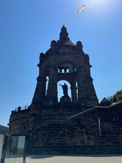 The image shows a low-angle, outdoor shot of a large stone monument against a clear blue sky. A paraglider with an orange and white wing is visible in the upper left corner, soaring against the bright sun. The monument itself is dark, constructed from rough-hewn stones and intricate brickwork, with arched openings and decorative elements. A dark statue of a person is visible within one of the large arches of the monument. The monument appears to be a historical or memorial structure, likely an outdoor attraction. The lighting suggests it is daytime, likely late morning or early afternoon, with the sun casting a strong light. The scene appears to be in a public space, possibly a park or historical site, given the monument's scale and the presence of railings and pathways. The orientation of the image is such that the monument appears to be tilted, suggesting the photo was taken with the camera held vertically and then rotated. The overall impression is one of grandeur and historical significance, with a hint of adventure provided by the paraglider. The location is identified as Porta Westfalica, Germany. There is no visible text in the image.
