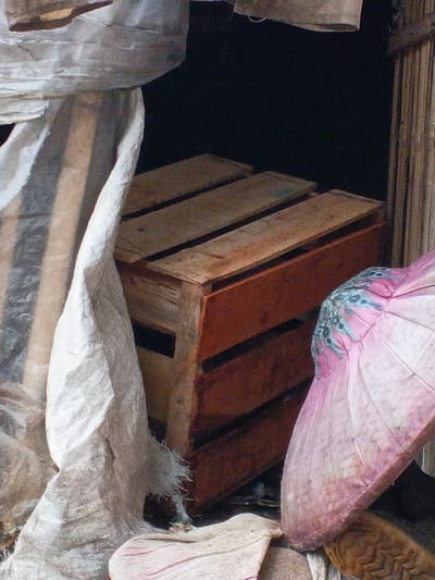 This image presents a static close-up view of several objects within a dimly lit, possibly indoor or sheltered space, in Welaluhu, East Timor.

The central subject is a wooden crate, constructed from rough planks. The top surface of the crate displays the natural, lighter wood grain, while the side panels are stained or painted a warm reddish-brown. This crate appears sturdy and utilitarian, perhaps used for storage or transport.

To the left, a substantial amount of light-colored fabric or sacking is draped and gathered, forming soft folds and curves. The material looks coarse and somewhat worn, suggesting its functional purpose.

On the right side of the frame, a distinctive pink object is visible. It has a structure reminiscent of a fan or a parasol, with radiating lines and a decorative section near the top featuring a pattern of blue and black shapes. This object introduces a vibrant splash of color against the otherwise muted palette of the scene.

The background is largely dark and indistinct, providing minimal context for the immediate surroundings. There are no people present, and no particular activity or event is depicted. The soft, diffuse lighting prevents the identification of the exact time of day but suggests an absence of direct, harsh sunlight. No text or specific location markers are visible on any of the items, apart from the general location context.