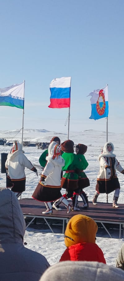 The image depicts an outdoor cultural performance in Nelmin-Nos, Russia, set against a vast, snow-covered tundra landscape under a clear blue sky. In the foreground, the backs of two spectators are partially visible; one wears a gray hooded jacket, the other a yellow knit hat and red jacket with black sunglasses resting on the back of the head. Six individuals perform on a dark, elevated stage in the midground. They wear traditional Indigenous clothing: white, fur-trimmed hooded parkas with geometric red, brown, and gold patterned ornamentation, dark lower garments, and patterned white and dark boots. Some also wear green tunics. Their dynamic poses suggest dancing or movement. The performer on the far right, wearing a white parka, faces forward and exhibits a smile. Behind the stage, three flags fly on poles: the flag of the Yamal-Nenets Autonomous Okrug, the Russian Federation flag, and the flag of the Nenets Autonomous Okrug. In the distant background, several vehicles, including a white SUV, and a herd of reindeer are present on the snow, with snow-covered hills on the horizon.