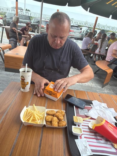 The image depicts a man sitting at a wooden table outdoors, likely at a McDonald's restaurant based on the branding visible on a cup and a food box. The man is wearing a dark t-shirt and is looking down at a small orange box in his hands, which appears to be a food container. In front of him on the table are a cup with a straw, a box of french fries, and a container of chicken nuggets. Also visible on the table are a smartphone and a tray with napkins and additional food items. In the background, other people are seated at tables, and cars are parked. The overall setting suggests a casual dining environment, potentially in a public space. The location is specified as Budykierz, Poland, with McDonald's branding indicating the specific establishment.