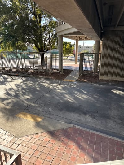 The image depicts an outdoor public area in Hialeah, United States of America, situated directly beneath a concrete elevated structure. The foreground features reddish-brown square brick pavers, which transition to a grey concrete and asphalt pavement. A rectangular yellow tactile paving strip is embedded within the concrete. Multiple, elongated shadows from trees and the elevated structure are cast across the ground, indicating strong overhead sunlight.

To the left, a grey metal bar fence separates the walkway from a green space. This fenced area contains a large deciduous tree, a palm tree, and at least two visible metal benches. A pole with a circular fixture is also present behind the fence. An opening in the fence, resembling a partially open gate, provides access to the green space.

The elevated structure is constructed from concrete beams and support pillars. A section of this structure, featuring a metal railing, is visible on an upper level to the left. On the immediate right, a vertically ribbed concrete wall forms part of a large pillar supporting the elevated pathway. In the background, beyond the fenced area, additional trees, several low-rise buildings, and utility poles are discernible under a clear sky. There are no individuals present in the scene.