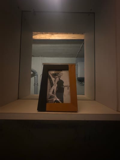 The photograph captures a framed black and white image of a woman in a dress and gloves, set against a dark backdrop. This frame, with its wooden and black elements, rests on a light-colored shelf. The shelf appears to be within a niche or recessed area, flanked by plain walls. Beyond the shelf and the framed picture, a view into another space is visible. This space reveals a concrete ceiling with visible wooden beams, exposed pipes, and a gray electrical box. The overall scene suggests an indoor setting, possibly a basement or utility area, located in Lapės, Lithuania.