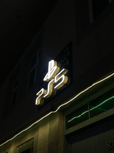 The image depicts the exterior of a building at night in Karmana, Uzbekistan. A prominent, illuminated white neon sign forming the "PS5" logo is mounted on the upper portion of a light-colored, textured building facade. To the left of this sign, a smaller, dark rectangular board featuring multiple white outlines resembling game controllers is visible. Below the main sign, a string of white LED rope lights extends horizontally across the facade, illuminating the surface directly beneath it. Below these lights, a row of dark windows reflects segments of green light. In the lower right corner, the building number "83" is faintly visible on the facade, adjacent to a dark, shuttered window or door. The scene is devoid of people.