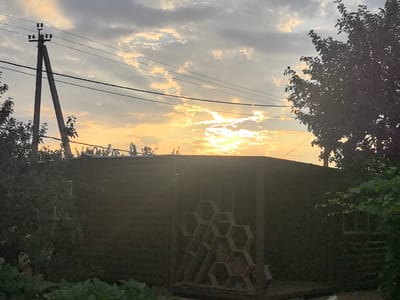 The image depicts a sunset over a rural landscape, possibly in or near the village of Perevoloki, Russia. In the foreground, a dark, low-slung building or shed dominates the lower half of the frame. To the left of the shed, lush green foliage and what appears to be a garden are visible. A unique hexagonal shelf unit is positioned against the side of the shed, holding several potted plants.
To the left of the building, a tall, angled utility pole with electrical wires stretches across the sky. On the right side of the image, the silhouette of a leafy tree frames the scene. The sky above is filled with a dramatic mix of clouds, illuminated by the warm, golden light of the setting sun, which is partially obscured by the clouds. The overall atmosphere is serene and indicative of a peaceful, rural evening.
