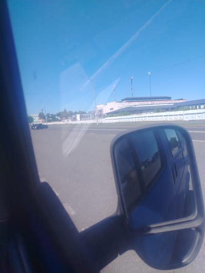 This is a medium-resolution, slightly blurry, wide-angle photograph taken from inside a vehicle, likely a car, looking out. The perspective is low, as if from the driver's or passenger's seat. The foreground is dominated by the dark silhouette of the vehicle's interior, casting a shadow across the lower left portion of the image. A side-view mirror, reflecting the blue exterior of the vehicle and the road behind, is prominently featured on the right side of the frame.
The scene outside the vehicle depicts a wide, paved road under a clear, bright blue sky. The time of day appears to be daytime, with the sun high and casting distinct shadows. The weather seems fair and sunny. In the mid-ground, a dark-colored car is driving on the road in the opposite direction. Further in the background, there are several buildings, some appearing to be industrial or commercial, with a fence in front of them. Multiple tall communication towers are visible against the sky, situated behind the buildings. The overall setting suggests an urban or suburban environment on the outskirts of a town or city.
The photograph lacks any visible text. There are no people clearly identifiable in the image, only the faint reflections in the side mirror which show what appears to be the interior of the vehicle. The image captures a fleeting moment of travel, with the focus on the road ahead and the reflection of the journey behind. The mood is neutral, simply documenting a scene from a moving vehicle.
