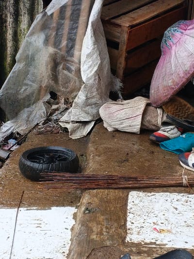 A cluttered outdoor scene reveals discarded items on a concrete surface. A black tire and a bundle of thin, brown sticks rest near worn-out shoes and a pink umbrella. Crumpled white and transparent plastic sheets drape over a wooden crate in the background. The area appears damp, with hints of trash scattered around. The composition suggests a neglected or storage space.
