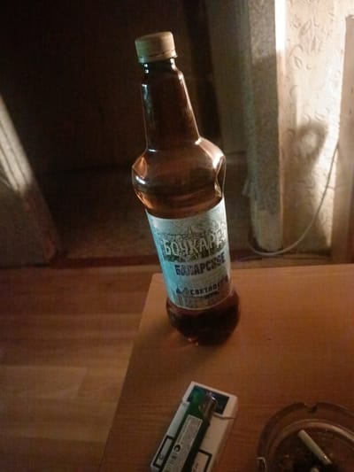 A bottle of "Bochkarev" beer, possibly light lager, is positioned at an angle on a wooden surface. The bottle has a greenish-brown hue and a light-colored cap. A white label with blue lettering is wrapped around the neck of the bottle, with the words "БОЧКАРЕВ" and "БАВАРСКОЕ" clearly visible. A white cigarette pack with a green lighter is placed on the same surface, to the right of the bottle. An ashtray containing cigarette butts is also present. The background is dimly lit and appears to be an interior setting, with a curtain visible on the right side. The scene suggests a casual setting, possibly a home or a bar. The image does not provide any direct information about the city of Voskresenskiy, Russia, as it is a general description of the objects and their arrangement.