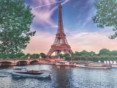 The image depicts a scenic view of the Eiffel Tower in Paris, France. The iconic landmark stands tall against a vibrant sky with streaks of pink and purple clouds and a blue gradient. In the foreground, a body of water, likely the Seine River, reflects the sky and the surrounding scenery. A white and grey passenger boat, with multiple people on deck, is cruising on the water, leaving a wake behind it. On the left side of the image, lush green trees frame the view, and on the right, a section of trees is also visible. In the mid-ground, a bridge with arched spans crosses the river, and along the riverbank, there are buildings, more trees, and a promenade populated by numerous individuals. Several other boats are docked along the riverbank near the Eiffel Tower. The scene captures a lively and picturesque Parisian atmosphere. The location is the city of Paris, France, not Kandyagash, Kazakhstan.