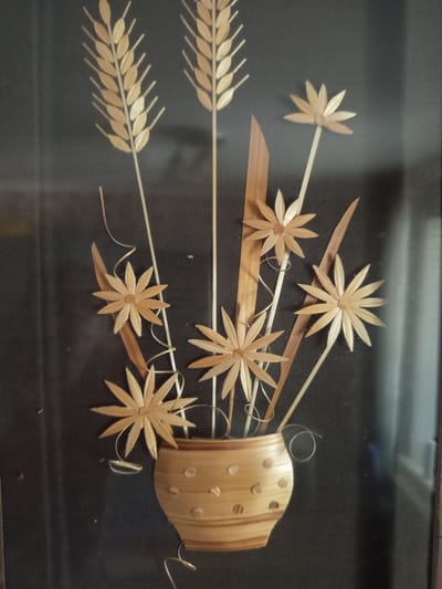This media file displays a decorative arrangement made from dried plant materials, likely wheat stalks and straw, mounted on a dark fabric background. The arrangement appears to be a still life, depicting a bouquet of stylized flowers and ears of wheat emerging from a vase-like structure.
The "flowers" are crafted from thin, pointed petals radiating from a central point, resembling daisies or sunflowers. There are five prominent flowers in the arrangement. Interspersed with the flowers are elements that resemble ears of wheat, with multiple small, pointed sections arranged along a central stalk. Two distinct ears of wheat are visible on the left side of the image.
The "vase" is a cylindrical, segmented object made from rolled or folded straw, with small circular indentations on its surface. Thin, white tendrils or strings are woven through the arrangement, adding a delicate touch. The overall composition suggests a rustic or folk art style.
There is no indication of people, a specific time of day, weather conditions, or any overt emotional expression. The background is uniformly dark, providing a stark contrast to the light-colored plant materials. The framing suggests the artwork is likely a framed piece, possibly for wall decoration. No visible text is present in the media file. The location context of Pavlohrad, Ukraine, suggests a possible origin for this type of folk craft, which is common in agricultural regions.