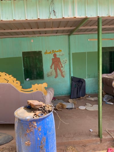 The image shows the interior of a building, likely a shop or a residence, located at latitude 15.552, longitude 32.57 in Khartoum, Sudan. The building has a green painted exterior and a brown roof. Inside, there is a large couch with a gold frame and a brown fabric upholstery. On the wall, there is a painted diagram of a human body labeled in Arabic with arrows pointing to different parts of the body. Next to the couch, there is a large blue barrel, possibly made of plastic, with rust marks on the surface. Several other objects, including a car part, are scattered on the floor, indicating a state of disarray or recent activity. There are no people present in the image.