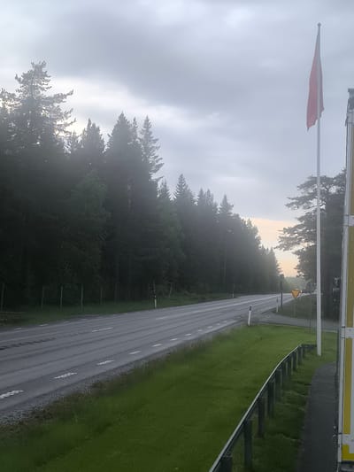 The image depicts a static outdoor scene in Bureå, Sweden, under an overcast and hazy sky, suggesting either early morning or late evening. A two-lane asphalt road, appearing damp, stretches diagonally from the lower left towards the upper right, gently curving into the distance. The road is marked with white dashed lane lines and solid white shoulder lines.
To the left of the road, a dense forest of coniferous trees, predominantly pines and spruces, extends throughout the background, partially obscured by atmospheric haze. A fence line composed of vertical wooden posts runs along the immediate edge of this forest.
On the right side of the road, a grassy verge is bordered by a dark metal guardrail. Beyond the guardrail, a paved area is visible, with a tall white flagpole holding a vertically hanging red flag that shows signs of fraying at its lower edge. Further right, a large yellow and white striped vertical surface, characteristic of a truck or building, is partially captured. In the mid-ground on the right, an intersection is indicated by a yellow triangular yield sign and pole-mounted traffic lights, flanked by more coniferous trees. No individuals or specific actions are discernible within the frame.