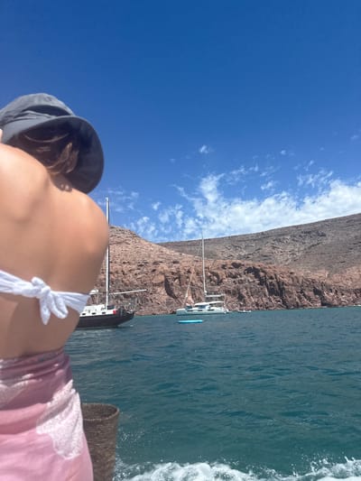 A woman with a hat and wearing a white bikini and pink cover-up is on a boat. The boat is traveling next to a rocky cliff. Other boats are in the water. The sky is blue with some clouds. The location is Unknown, Mexico.