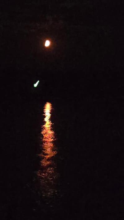 The image depicts a nighttime scene over a body of water, likely a river or lake, in the Жаңабұлақ ауылдық округі region of Kazakhstan. The sky is dark, with a partially illuminated moon visible in the upper left quadrant, casting a soft orange glow. Below the moon, a small, green light source is present, possibly a navigation marker or a distant light. A reflection of an orange-red light, likely from the moon or another external source, shimmers on the water's surface, creating a streak of light that extends downwards. The surrounding environment is predominantly dark, with the textures of the water and the faint atmospheric conditions being the only visible elements besides the light sources. No people or specific structures are identifiable within the frame, suggesting a natural or sparsely populated setting at night.