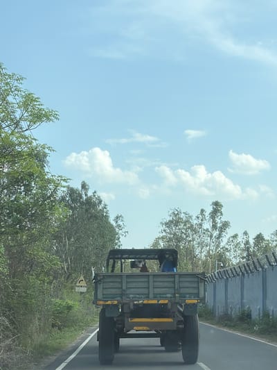 In Pudupakkam, India, a green military-style truck with a flatbed drives on a paved road. Two people are visible in the truck's cab, one wearing a blue shirt. The truck is traveling on a road bordered by lush green vegetation on the left and a grey concrete wall topped with barbed wire on the right. The wall appears to be a security fence. The sky is a clear blue with scattered white clouds. A yellow and black directional sign pointing right is visible on the left side of the road, amidst the trees.