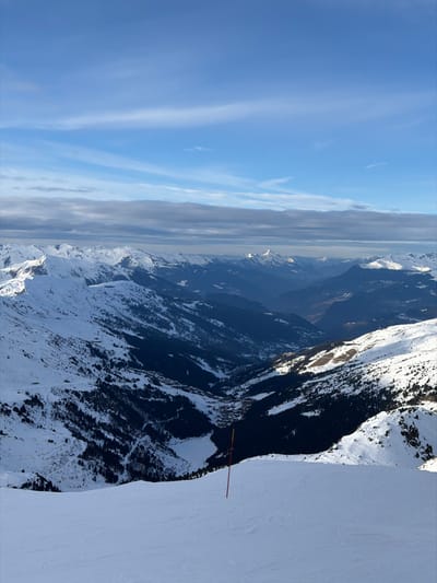 Mountains covered in snow reach into a blue sky with streaky clouds. A valley stretches into the distance, partially obscured by haze. A steep, snow-covered slope dominates the right foreground, with a warning sign marking the edge. The mountain's rugged terrain is visible through patches of trees and snow.