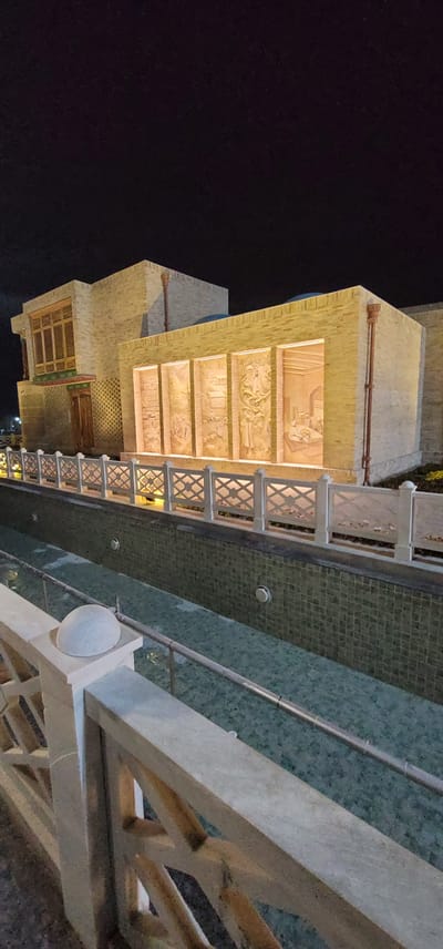 The image shows a building exterior at night in Satkak, Uzbekistan. The building is constructed of light-colored bricks and features decorative relief panels illuminated from behind. A white ornate fence runs along the front of the building, separating it from a shallow, tiled water channel. The water channel has a few circular fixtures visible within it. The sky is dark, indicating it is nighttime. No people are visible in the image.
