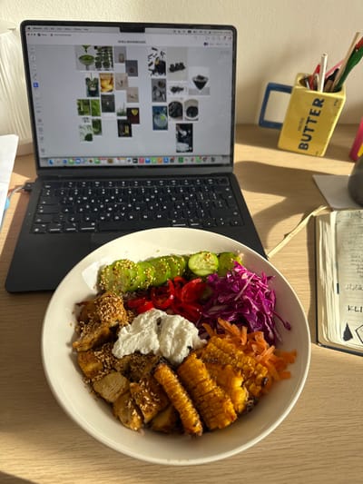 This image captures an indoor scene on a light-colored wooden desk, located within the Anavissos Municipal Unit, Greece. A black MacBook laptop, open and displaying an "AVRA MOODBOARD" with various design images, is positioned centrally. Its keyboard, featuring a localized layout with Greek characters, is partially visible. In front of the laptop, a white ceramic bowl contains a colorful meal. The meal includes sections of sliced cucumbers with sesame seeds, thinly sliced red peppers, shredded purple cabbage, shredded orange carrots, roasted corn on the cob segments, and cubed protein (likely chicken or tofu) also topped with sesame seeds, alongside a dollop of white creamy substance. To the right of the laptop, a yellow ceramic stationery holder shaped like a stick of "SALTED BUTTER" holds multiple pens and pencils. Adjacent to the stationery holder, a dark blue or black hardcover notebook with handwritten pages and a light-colored ribbon bookmark is visible. Strong natural light enters from the left, casting distinct shadows from the objects onto the desk surface. No individuals are present in the scene.