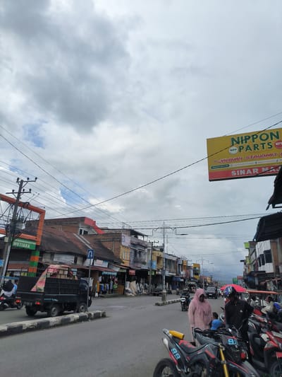 The image depicts a street scene in the city of Mutiara, Indonesia. The foreground features several motorcycles parked on the side of the road, with one prominently displaying the number 99. Two people, one wearing a pink headscarf and the other a helmet, are standing near the motorcycles, appearing to interact. Further into the scene, a black pickup truck is being driven away from the viewer, carrying what appears to be bedding or fabric on its bed. On the right side of the street, buildings with storefronts line the road, displaying various signs, including one for "NIPPON PARTS" with the tagline "JAMINAN STANDART" and "SINAR MA". On the left side of the street, there is a utility pole with numerous electrical wires crisscrossing the sky. The sky is overcast with large, grey clouds. The road is paved, and there are sidewalks and curbs visible. The overall atmosphere suggests a busy urban environment with commercial activity.