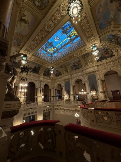 The image depicts the opulent interior of the Grand Hotel in San Pellegrino Terme, Italy. The photograph captures a grand staircase and atrium, looking upwards towards a stained-glass ceiling. Ornate architectural details, including elaborate moldings, stucco work, and painted murals adorn the walls and ceilings. Several large, decorative chandeliers hang from the ceiling, illuminated with warm light. A prominent statue of a muscular male figure is visible on the left side of the frame, positioned on a pedestal. The balconies and railings of the staircase are also elaborately decorated, with red velvet upholstery on the top surfaces. The overall impression is one of grandeur and historical opulence, characteristic of Belle Époque architecture.