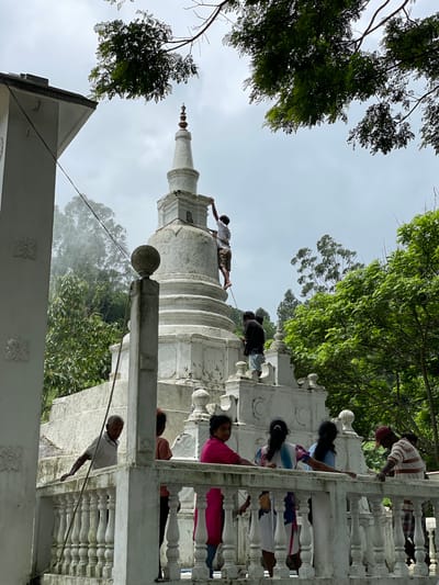 The image shows a group of people cleaning a white stupa in Kumbalwela, Sri Lanka. One man is atop the stupa, while others are on the building's railings, using hoses and cloths. The stupa is surrounded by trees and under a cloudy sky. The architecture features white structures with detailed railings. The people appear to be engaged in a cleaning or maintenance task.
