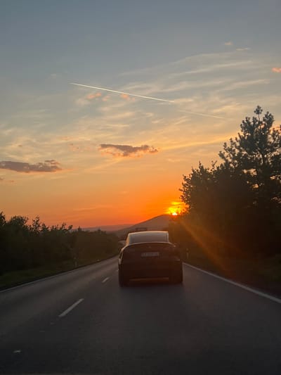 The image depicts a sunset scene alongside a road. A dark-colored car is parked on the shoulder of the road. The sky transitions from light blue to orange with some clouds and a contrail. Trees line the side of the road near the sunset. The license plate on the car is visible, showing 'EA 2595 AE.' The location is Kesarevo, Bulgaria.