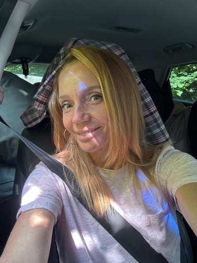 A woman with blonde hair and a pink t-shirt is sitting in the passenger seat of a car. Sunlight is shining through the window, casting a blue and white reflection on her face and shirt. She is wearing hoop earrings and has a seatbelt fastened across her chest. A plaid blanket is draped over her head. The background shows the interior of the car and greenery outside the window. The scene takes place in Portovoe, Russia.
